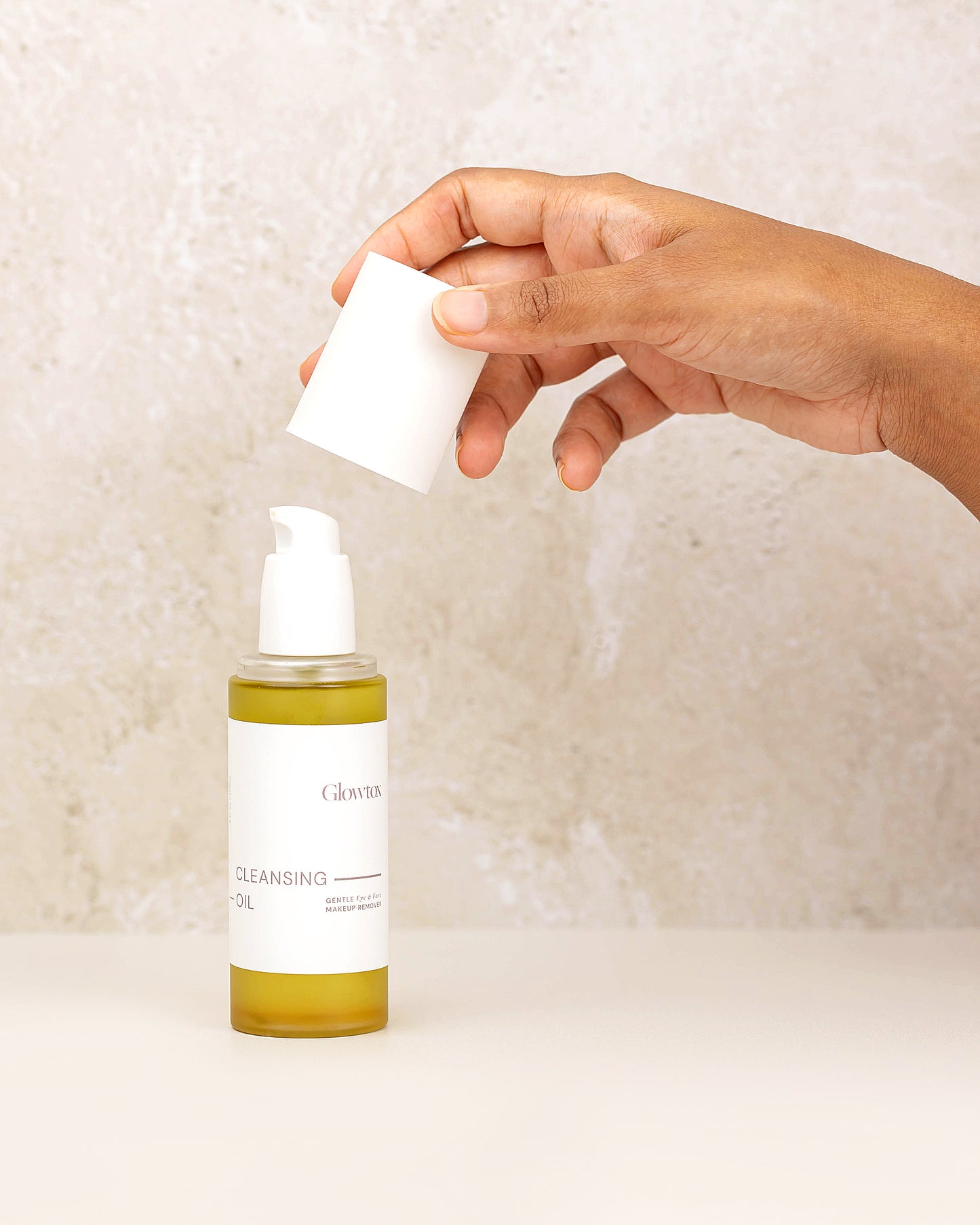 A hand lifts a white cap off a bottle of Glow Recipe cleansing oil, set against a textured beige background.